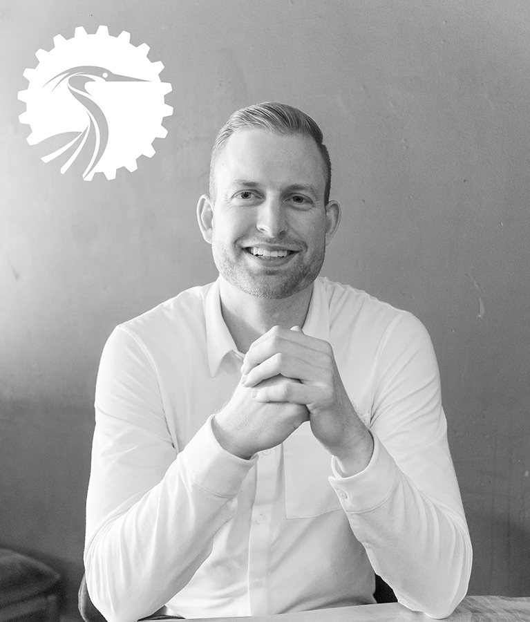 A man with short hair sits at a table, smiling, with his hands clasped. He wears a light-colored shirt. A manufacturing logo featuring a heron inside a gear is visible on the wall behind him. The image is in black and white.