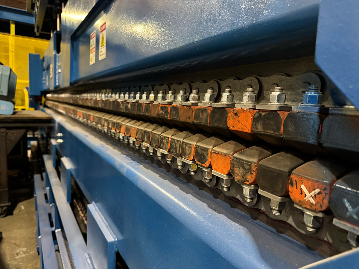 Close-up view of an industrial machine with a row of metal bolts and angular, interlocking components, some marked with orange paint. The machine is blue, and there are warning labels visible.