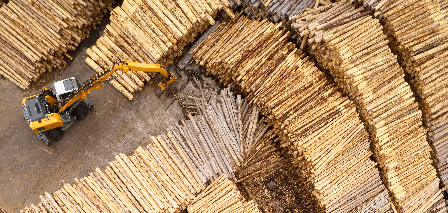 Aerial view of a yellow excavator stacking large piles of logs at a Forestry Services lumberyard. The logs are neatly arranged in long rows, showcasing various shades of brown and beige.