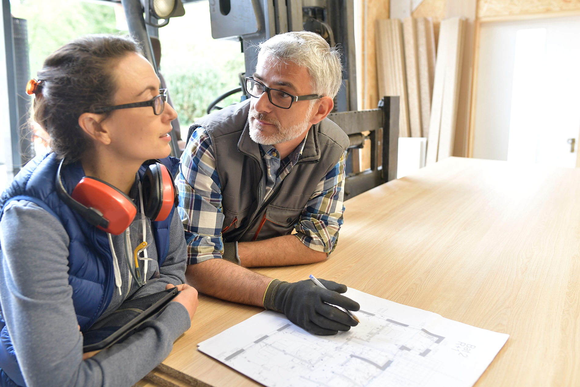 Two people in a workshop discuss blueprints on a wooden table; one wears glasses and gloves, the other has headphones around her neck. Both appear engaged in conversation about woodworking materials and Forestry Services.